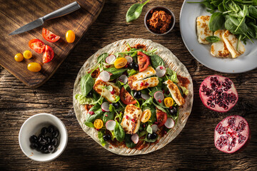 Top view of an open top vegetarian tortilla with salad, raddish, chery tomatoes, olives, pomegranate and grilled haloumi cheese