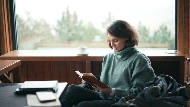 A young cheerful woman sitting in her living room with panoramic windows and a view of the forest, reading a book and drinking coffee from a cup