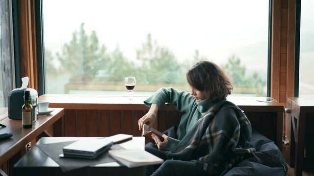 A young cheerful woman sitting in her living room with panoramic windows and a view of the forest, reading a book and drinking red wine from a glass.