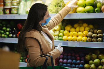Happy woman in protective mask taking fresh vegetables while standing by groceries in supermarket. Beautiful young girl with food basket choosing food by stand with vegetables. Christmas purchases
