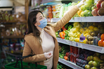 Happy woman in protective mask taking fresh vegetables while standing by groceries in supermarket. Beautiful young girl with food basket choosing food by stand with vegetables. Christmas purchases