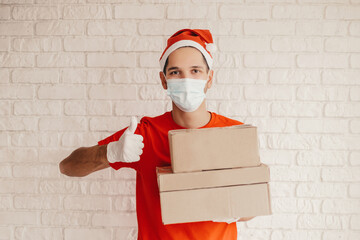 Happy man courier in protective face mask, medical gloves gesture ok sign and smile, hold cardboard boxes in hand. Young delivery man in santa hat, face mask carry packages. Winter holidays delivery