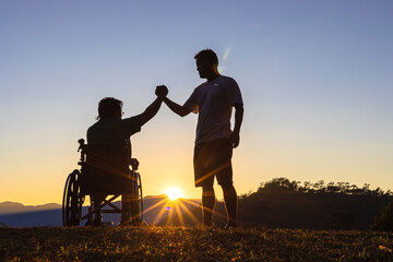 Fototapeta na wymiar Silhouette of joyful disabled man in wheelchair raised hands with friend at sunset