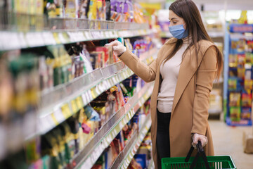 Horizontal shot of woman in protective mask and gloves reading label with price at goods while standing in supermarket. Beautiful young girl choosing snaks while doing shopping at grocery shop