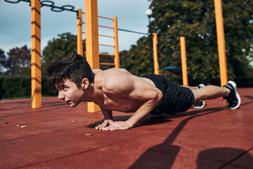 Young shirtless man bodybuilder doing push-ups on a red rubber ground during his workout in a modern calisthenics street workout park