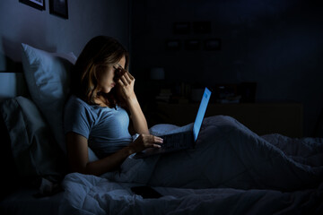Young woman having sore and tired eyes when using laptop while sitting in bed at night