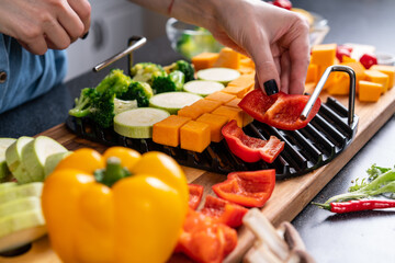 Female hand lays out chopped vegetables on grill grate, in home kitchen. Blured foreground.