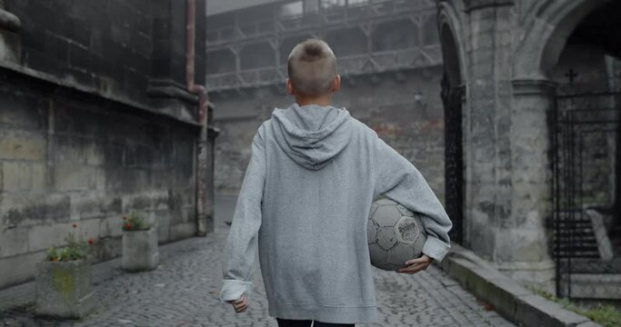 Crop view of teen guy in casual clothes holding football ball.Backside view of boy walking at old european city street. Concept of sport, leisure, lifestyle, childhood. Outdoors