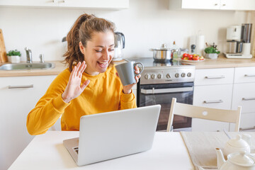 Smiling girl waving hand video calling family by webcam. Woman with laptop having virtual meeting chat video call conference sitting on kitchen at home. New normal social distance self isolation.