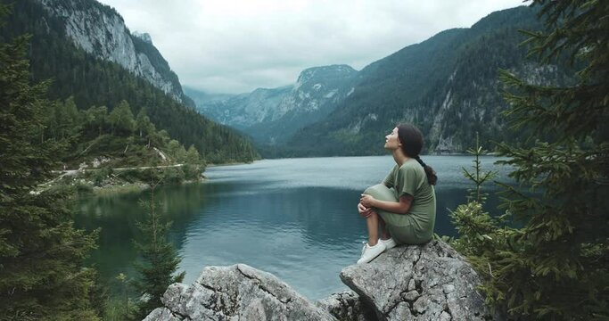 Beautiful calm woman sitting on rock coastline. Girl having moment alone, uniting with nature and enjoying life. Magnifficent background of clear lake in mountains. Concept of travel, inspiration.