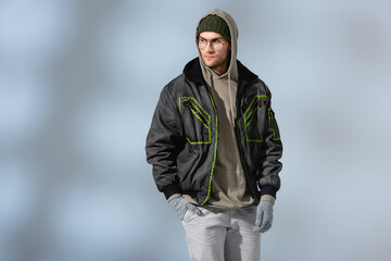 fashionable man in glasses, hat, anorak and gloves standing with hand in pocket on grey © LIGHTFIELD STUDIOS