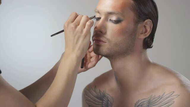 Tilt up shot of male MUA using brush and blending eyeshadow on shirtless young man