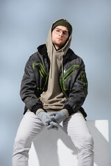 young trendy man in hat and anorak sitting on white cube with clenched hands in gloves on grey