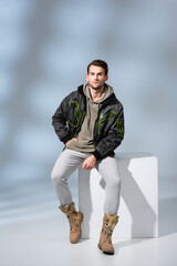 full length of trendy man in boots, hoodie and parka posing while sitting on white cube on grey © LIGHTFIELD STUDIOS