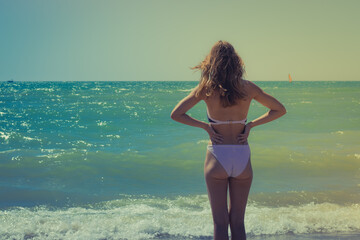 Skinny girl in a swimsuit, the view from the back, stands by the sea and prepares to swim. Water background and copy space