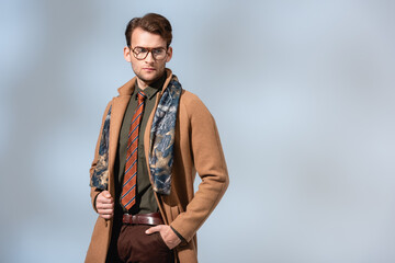 stylish man in glasses and winter coat standing with hand in pocket on grey