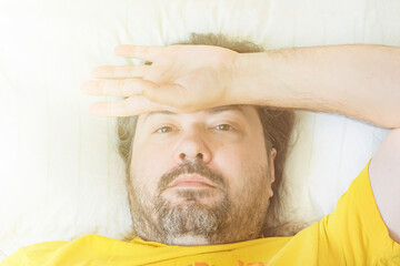 A middle-aged bearded man lies on a pillow in the early morning with his hand on his forehead. Concept - Early Awakening at Work