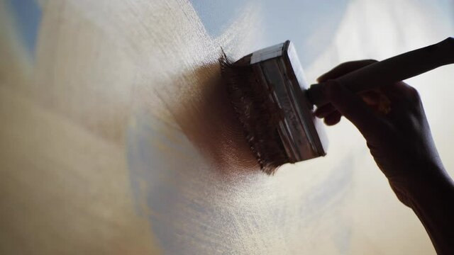 Close-up of artist holding paint wide brush and drawing oil painting at the studio. Unusual new abstract painting, brown, gold line on light canvas. One example of a painting technique.