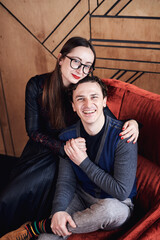 Young woman in eyeglasses and man sitting on red velvet Santa armchair in front of wooden wall, smiling, laughing. Romantic relationship. Close-up picture of young couple in fancy clothes.