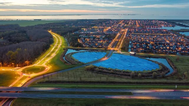 Modern sustainable residential neighbourhood in the city of Almere, The Netherlands, with solar panels farm powering city heating. Evening traffic on the roads. Aerial Hyperlapse shot at dusk. 