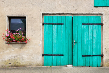 Fototapeta na wymiar Frontal closeup view of a weathered beige farmhouse with turquoise green hinged barn doors and window with a flowerpot on the facade, in the German countryside