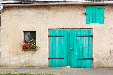 Fototapeta na wymiar Frontal wide view of a weathered beige farmhouse with turquoise green hinged barn doors and window with a flowerpot on the facade, in the German countryside
