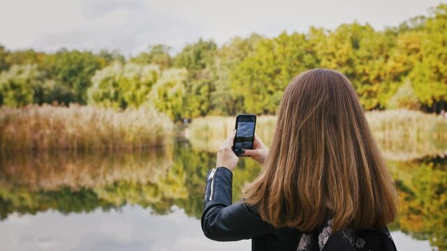 Young woman with backpack taking photos of autumn landscape, capturing lake and forest nature on smartphone, back view