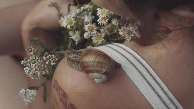 Close-up of snail slowly crawling along shoulder of young girl. Woman holding bouquet of wild flowers or daisies in her hands. Snail leaves muscus on skin. Concept of nature, cosmetology, spa, beauty.