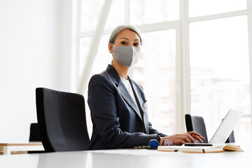 Focused white-haired businesswoman in face mask working with laptop