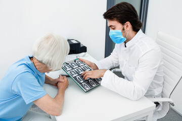 Fototapeta na wymiar Pulmonologist wearing a surgery mask showing a senior patient a CT scan of her lungs. Pneumonia, coronavirus, lung disease