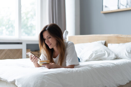 Beautiful young woman relaxing on bed at home, using mobile phone, holding credit card, shopping.