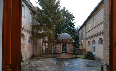 Chiostro ex monastero con pozzo visto da dietro ai vetri di una grande porta finestra 