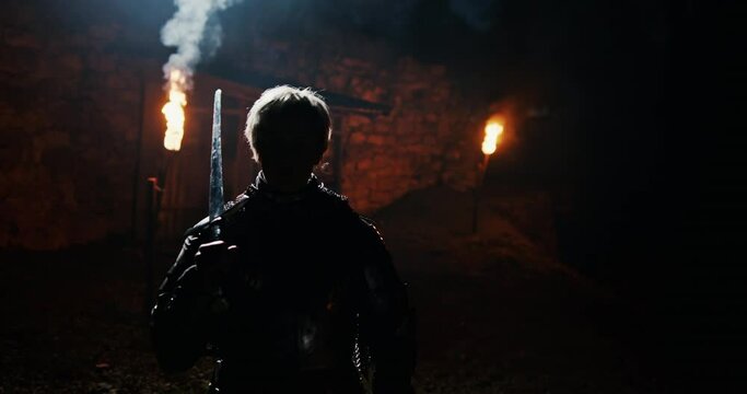 Anonymous female warrior examining sword. Unrecognizable woman in Joan of Arc armor touching and inspecting sword against house with torches at night