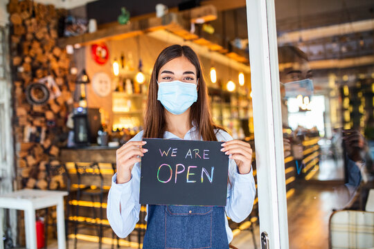 Happy female waitress with protective face mask holding open sign while standing at cafe or restaurant doorway, open again after lock down due to outbreak of coronavirus covid-19