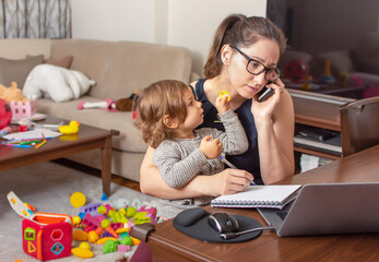 Young beautiful business woman talking on mobile phone and working on a laptop. Mother playing with...