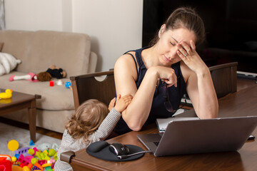 Tired mother trying to work on a laptop at home during her kid crying. Childcare and working mom concept. Women powerful. Toddler tantrum. Young lady working at home during quarantine.