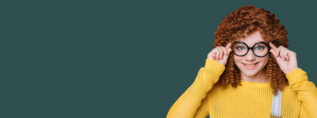 Cheerful red-haired curly woman holding trendy glasses near her face, isolated over tidewater green...