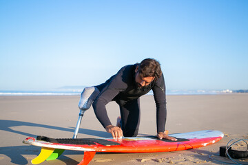 Sporty man in wetsuit wearing artificial limb, waxing surfboard on sand on ocean beach. Artificial limb and active lifestyle concept