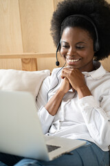 Smiling Afro-American millennial woman wearing headset, communicating via video chat on laptop,...