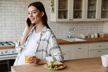 Happy pregnant woman talking on cellphone while having breakfast