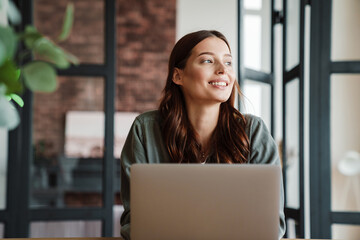 Beautiful smiling woman working with laptop while sitting at table - Powered by Adobe