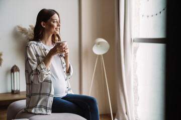 Beautiful happy pregnant woman smiling and drinking coffee while sitting on couch