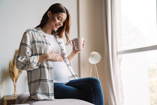 Beautiful happy pregnant woman smiling and drinking coffee while sitting on couch