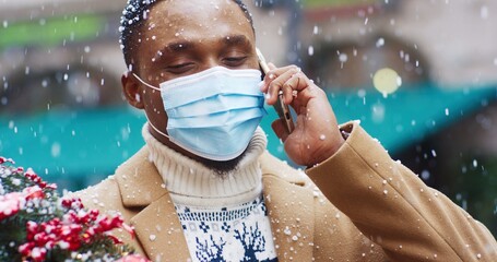 Handsome African American man in medical mask stands with Christmas tree in his hands and talks on cell phone under falling snow.