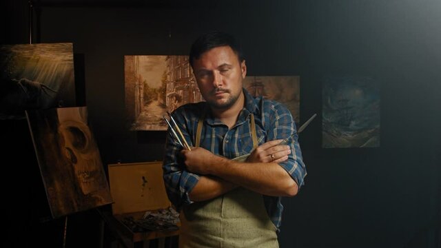 Portrait of the talanted artist with spatula and paintbrushes in his hands after finishing the new masterpiece of the oil painting. Fine art concept