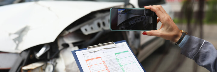 Woman photographs a broken car on smartphone and holds insurance documents in her hands. Damage...