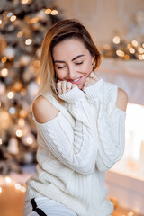 Portrait of a pretty blonde with a cozy home sweater at the Christmas tree, waiting for the new...