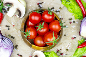 Top view of fresh red ripe cherry tomatoes in wooden bowl with salad leaves, onion, pepper, champignon mushrooms, spices and salt on light food background.