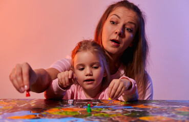 Fototapeta premium The family is having fun playing together at home in a board game. Board game field, many figures, coins and sandglass. Mother and cute little daughter play, holding cards joyful emotions on the face.