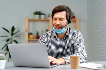 Young man working on laptop with headset and face mask at home